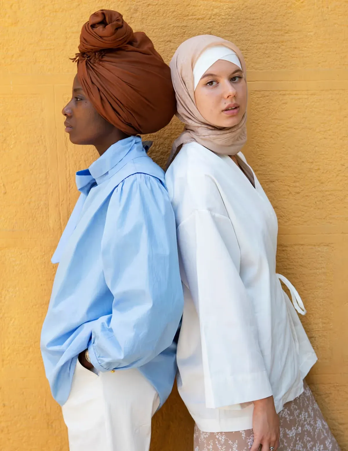 Two women back to back against a yellow wall, one in a rust head wrap, one in a beige hijab