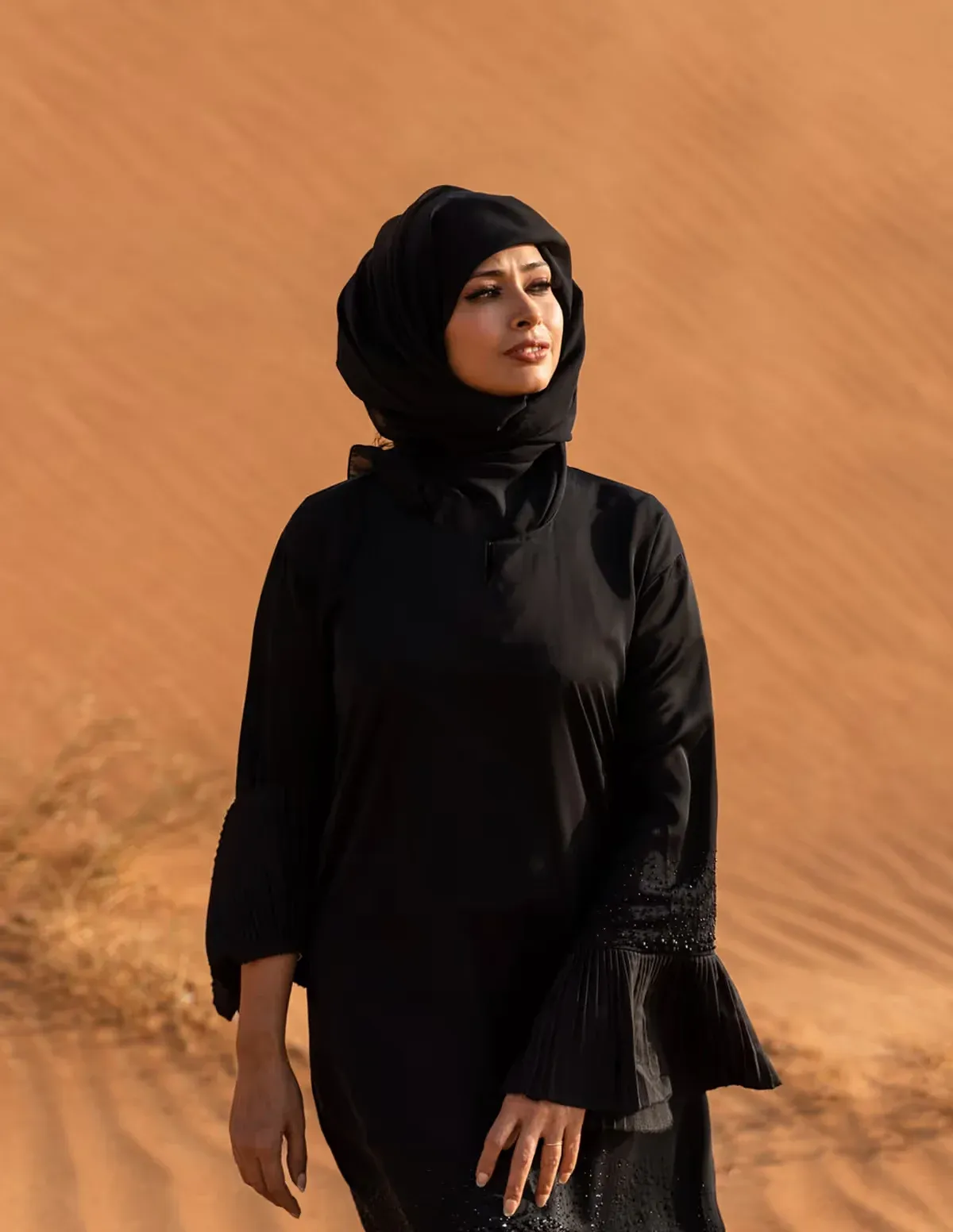A woman in a black hijab and long black tunic walking on desert sand
